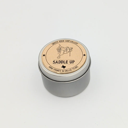 Silver metal candle tin labeled “Saddle Up” with a western saddle illustration, hand-poured in Dallas, Texas, photographed on a clean white background.