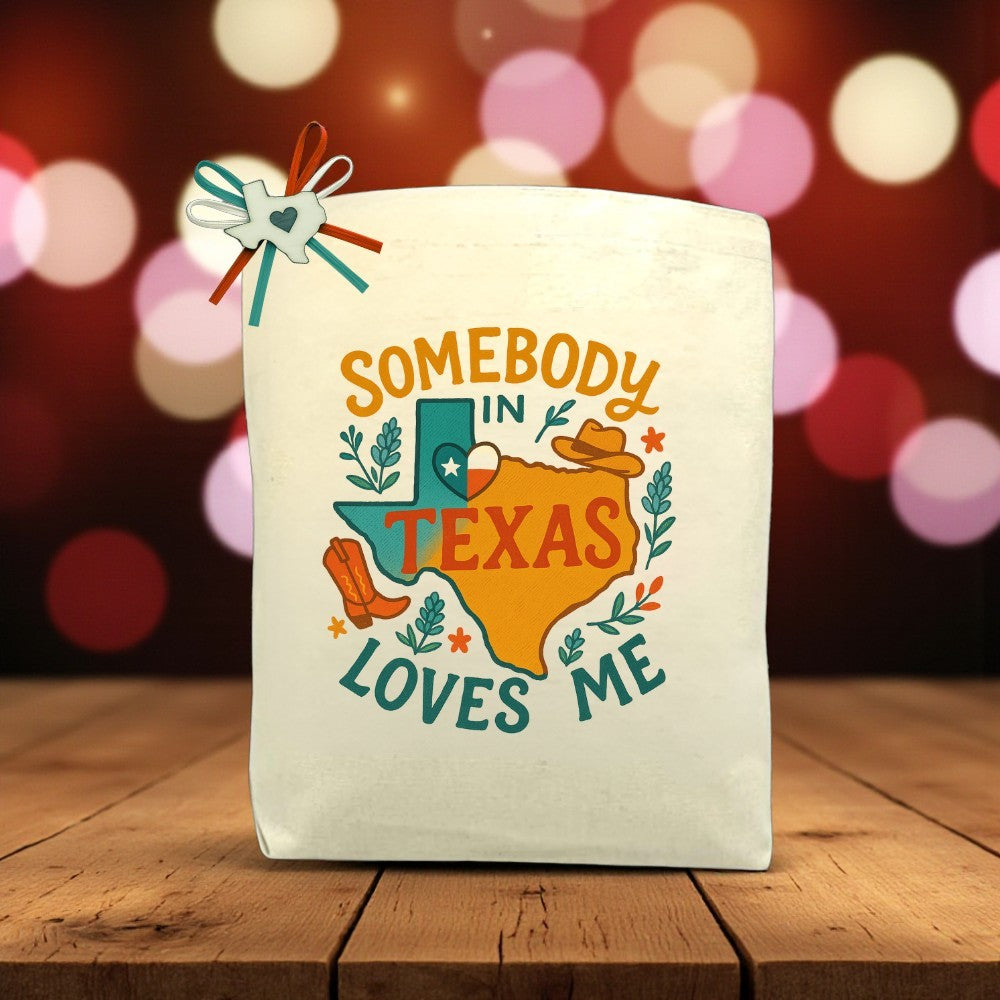 Somebody In Texas Loves Me canvas tote bag with Texas-themed design, cowboy hat, and boots, displayed on a wooden table
