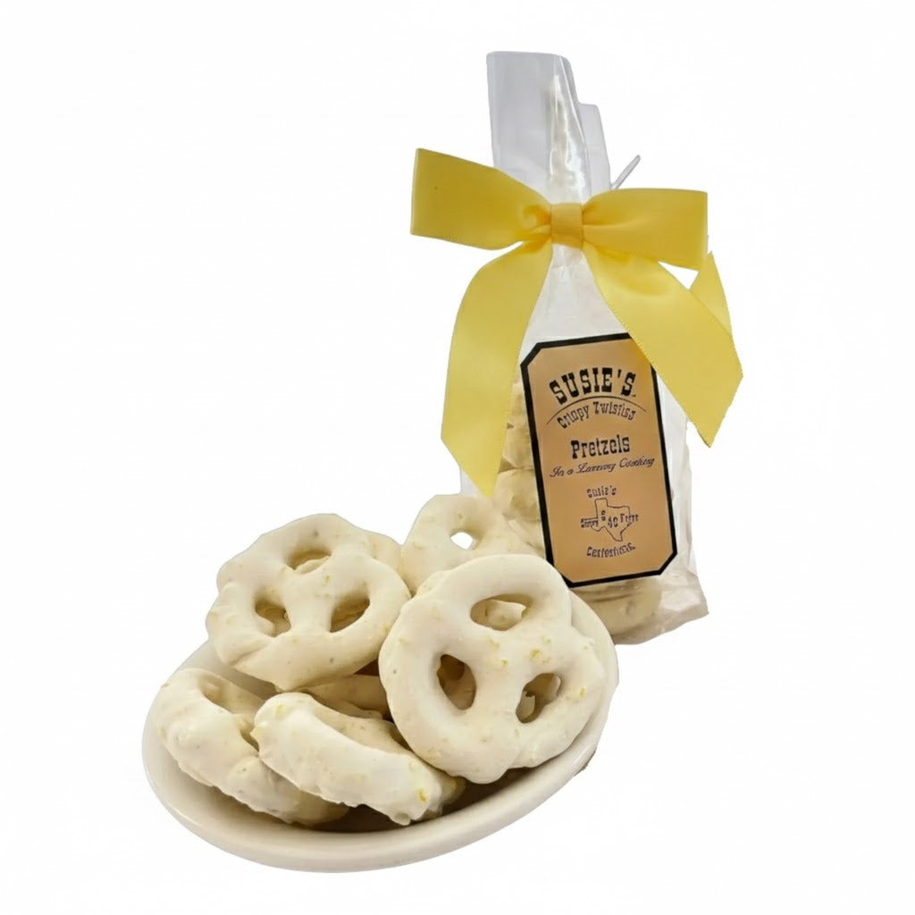 White chocolate–coated pretzels on a small dish beside a clear gift bag of Susie’s Crispy Twisting Pretzels tied with a yellow ribbon, photographed on a white background