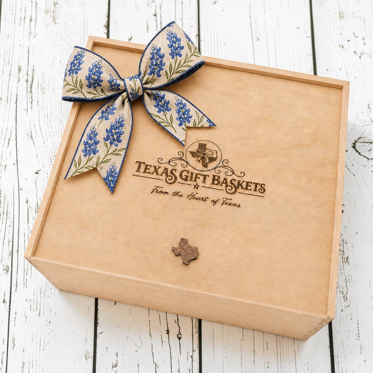 A square kraft paper gift box with a large blue floral ribbon bow on top, photographed on a white rustic wood surface. The box is printed with the Texas Gift Baskets logo and a small Texas-shaped ornament on the front.
