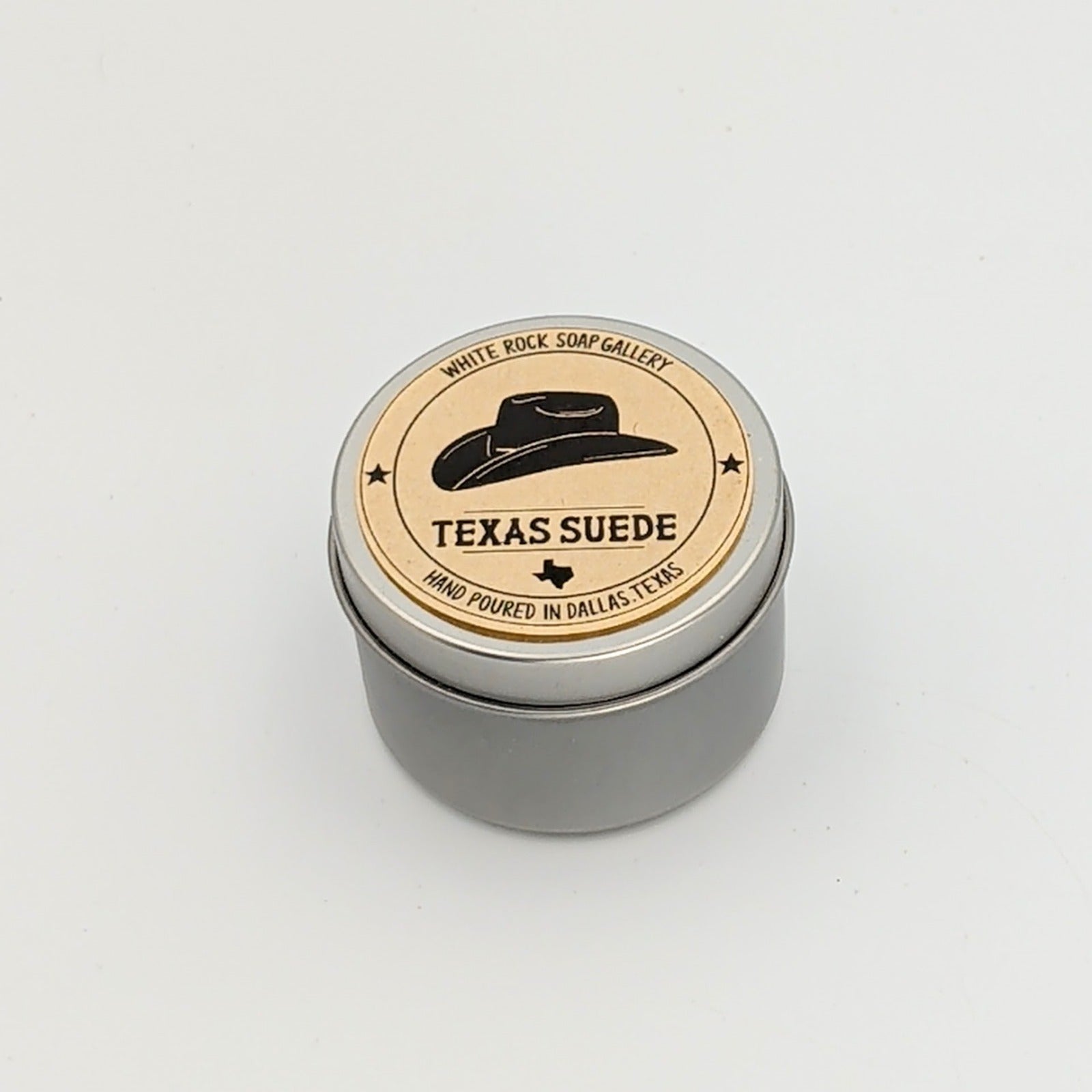 Round silver tin candle labeled “Texas Suede” from White Rock Soap Gallery, featuring a tan label with a black cowboy hat graphic and text noting it is hand poured in Dallas, Texas, displayed on a white background.
